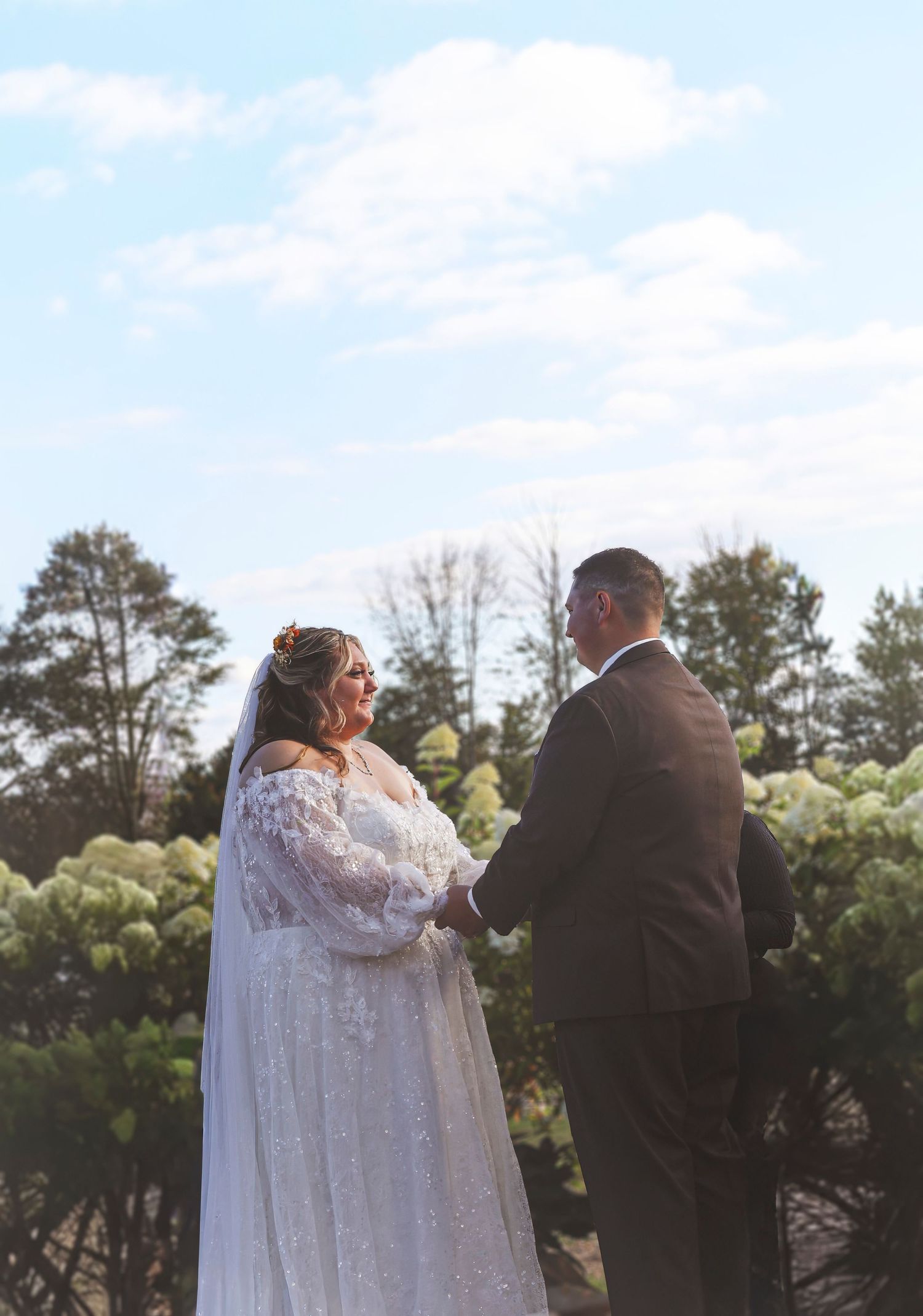 Bride in lace gown and veil holds hands with groom in suit during outdoor wedding ceremony under blue sky.