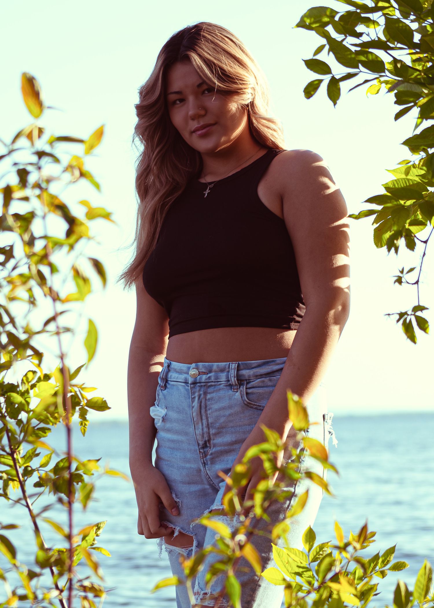 Young woman in black crop top and ripped jeans posing among green leaves with ocean in background.