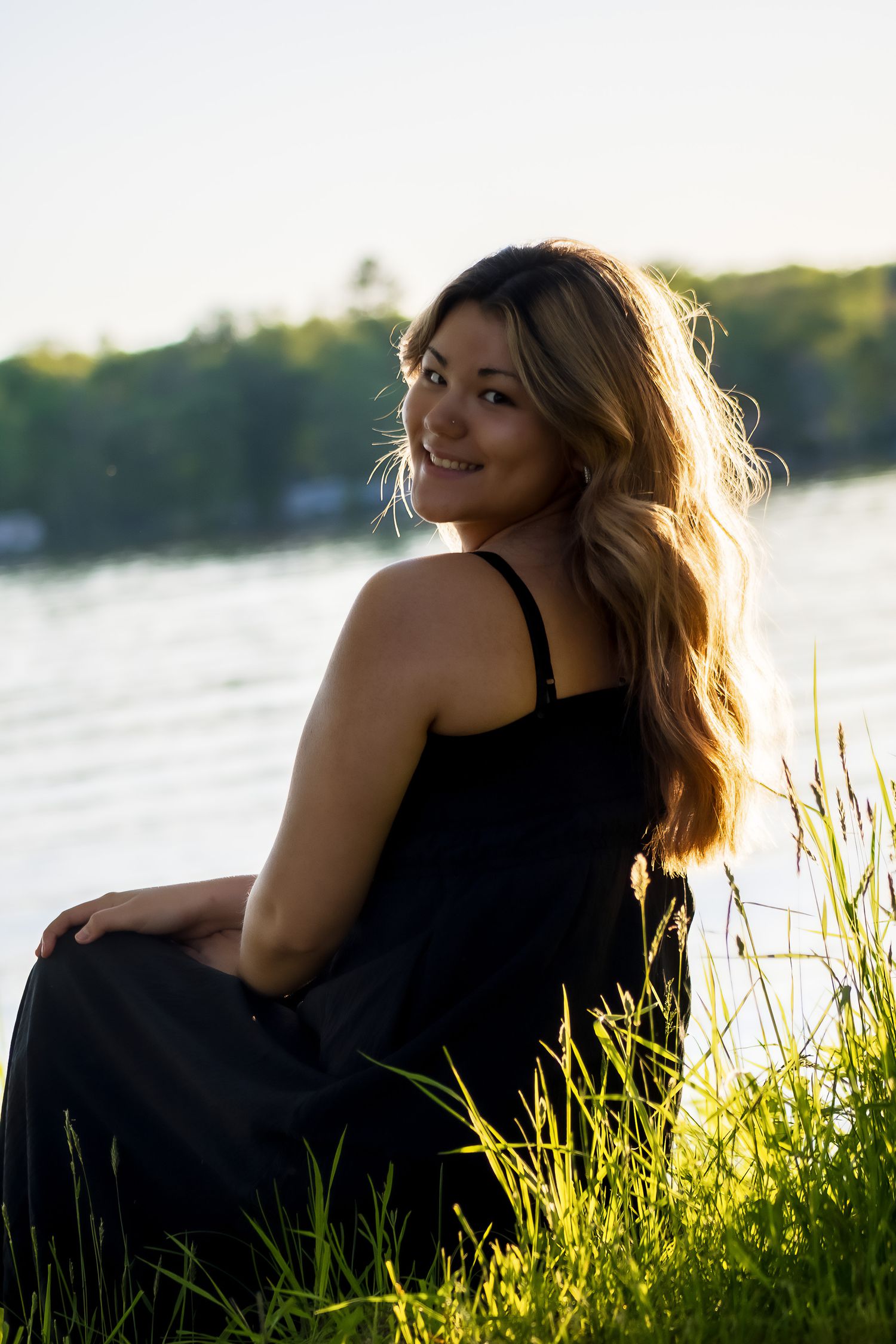 Young woman in black dress sitting on grass by a sunlit lake, smiling over her shoulder with long flowing hair.