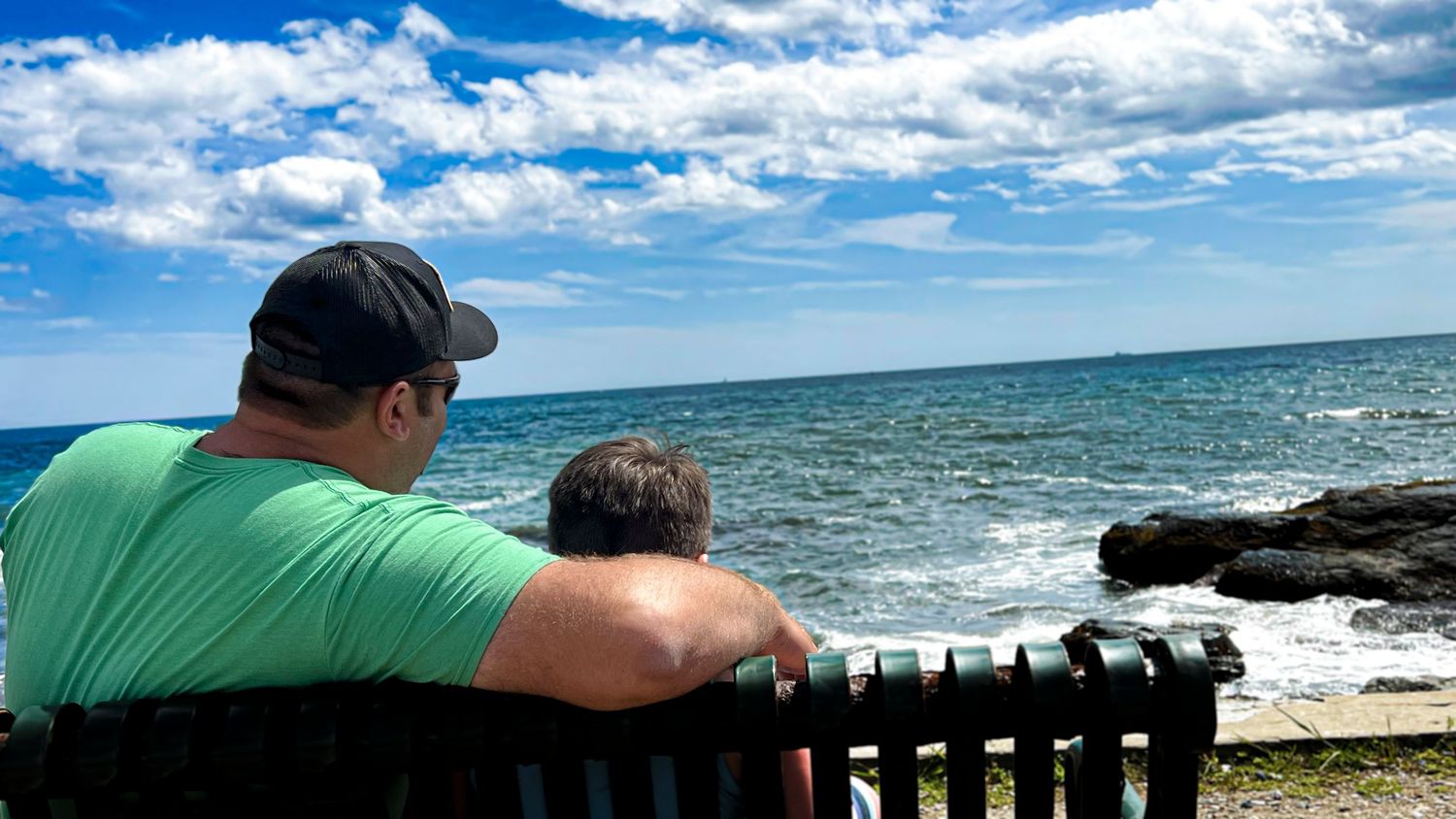 Two people sit together on a bench overlooking a rocky ocean shoreline under a bright blue sky.