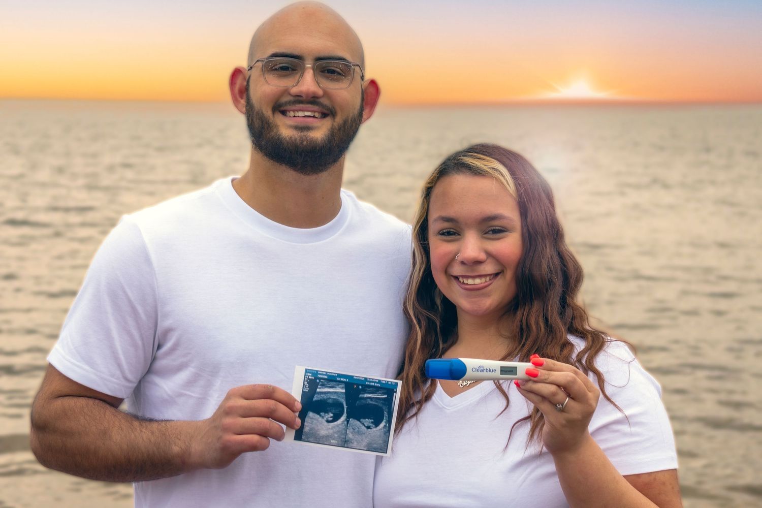 Couple in white shirts holding ultrasound photos and pregnancy test on beach at sunset.
