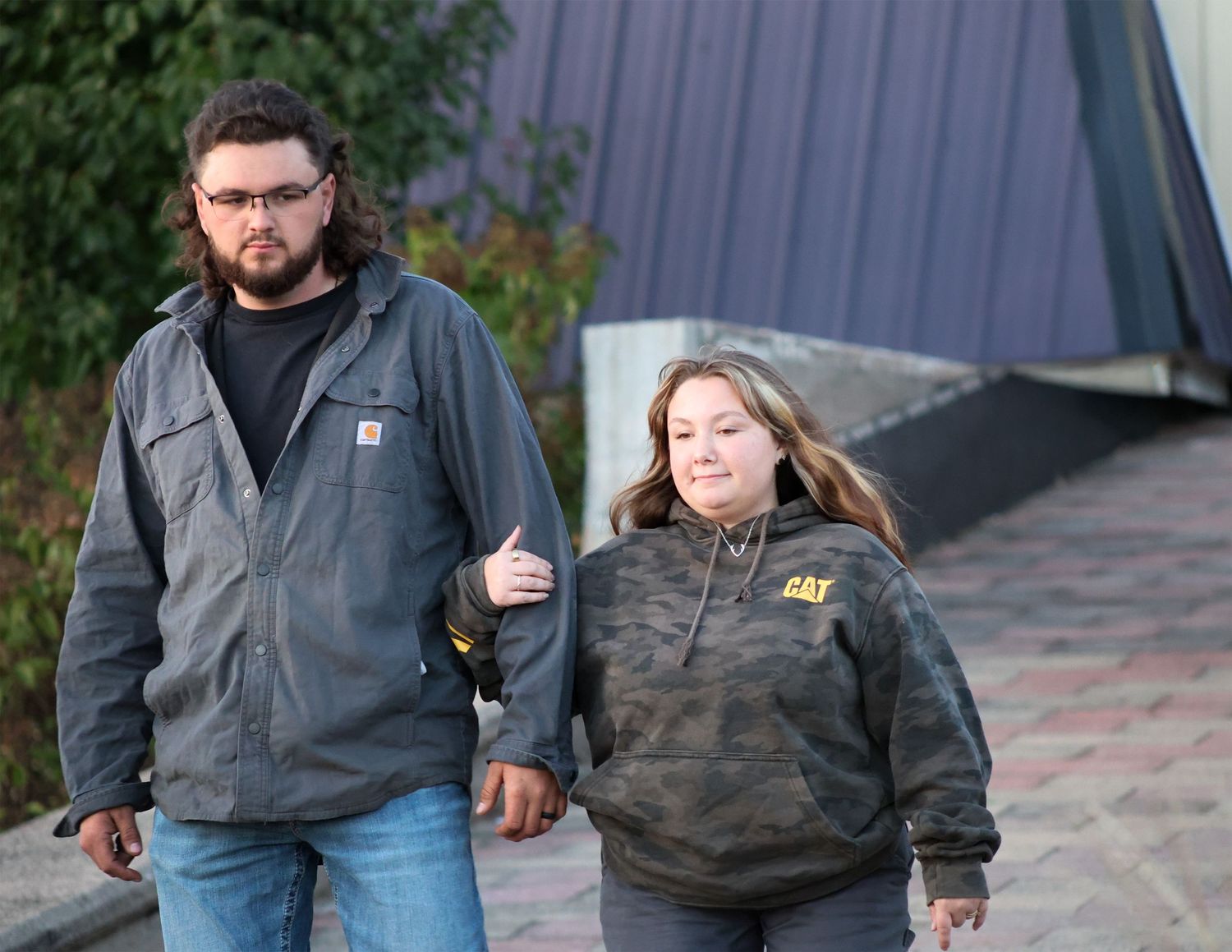 A man and woman walking together outdoors, wearing casual jackets, on a paved pathway near a building.