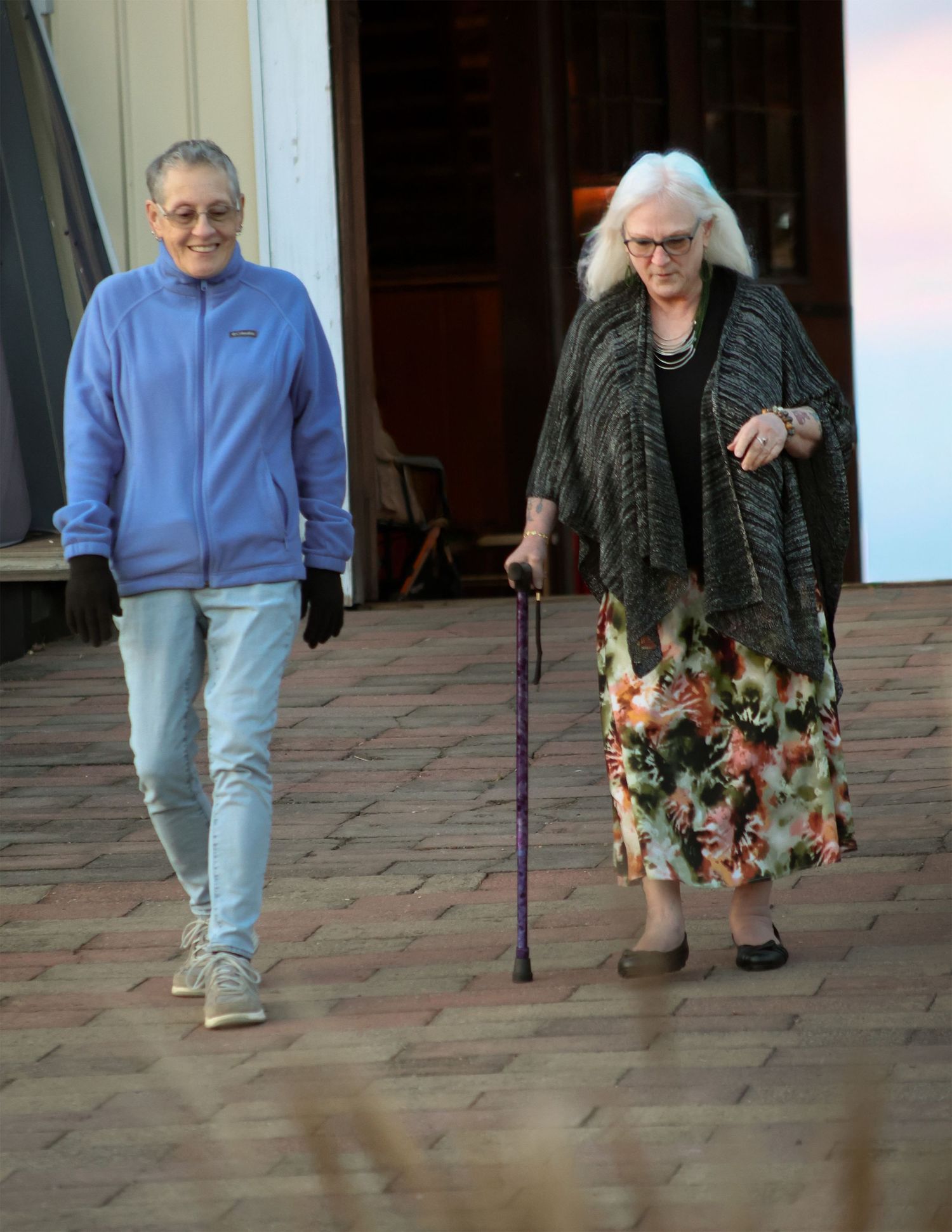 Two women walking on brick path, one in blue fleece jacket and jeans, other in floral skirt using purple cane.