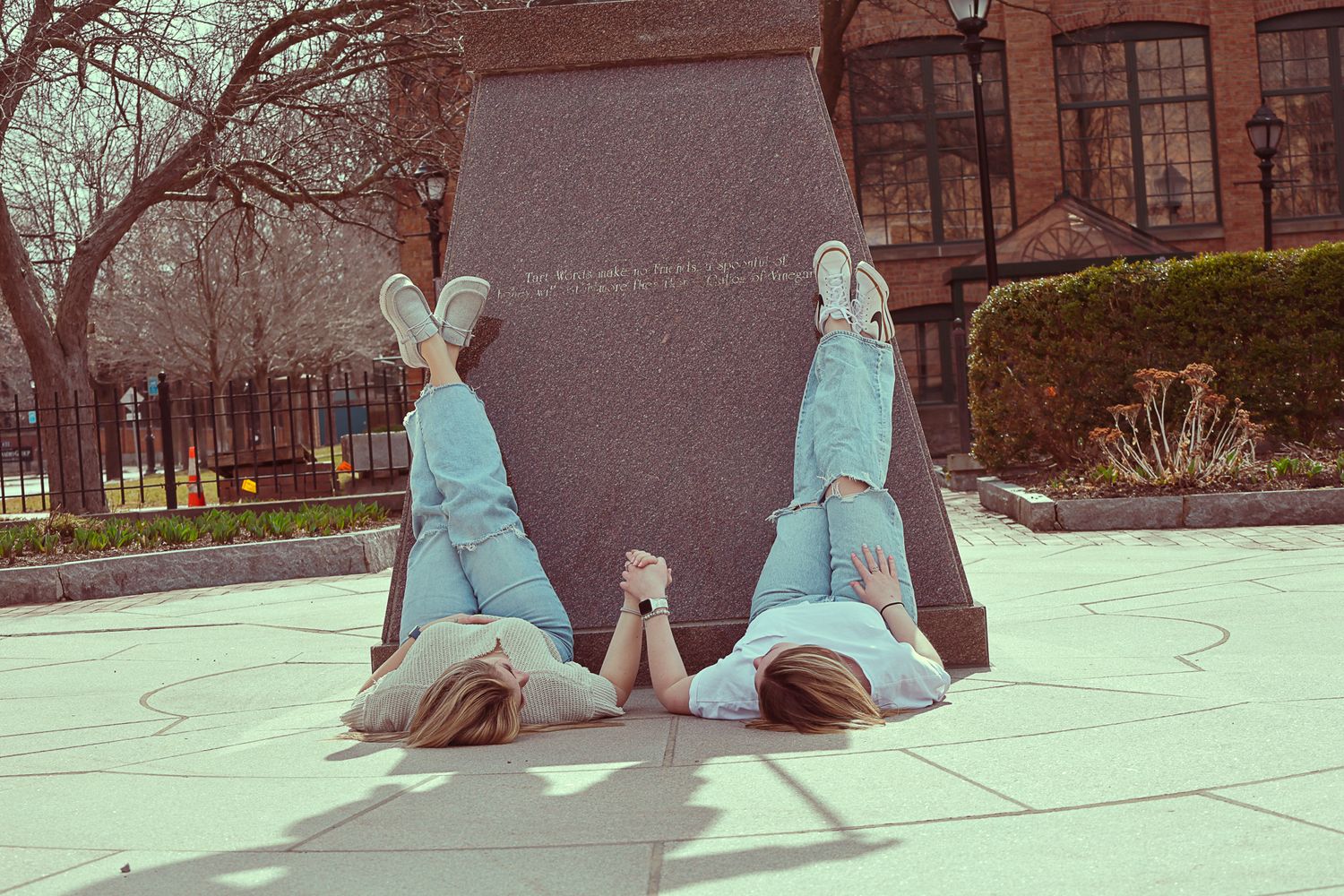 Two girls lying on ground with legs up against a large black monument outdoors.