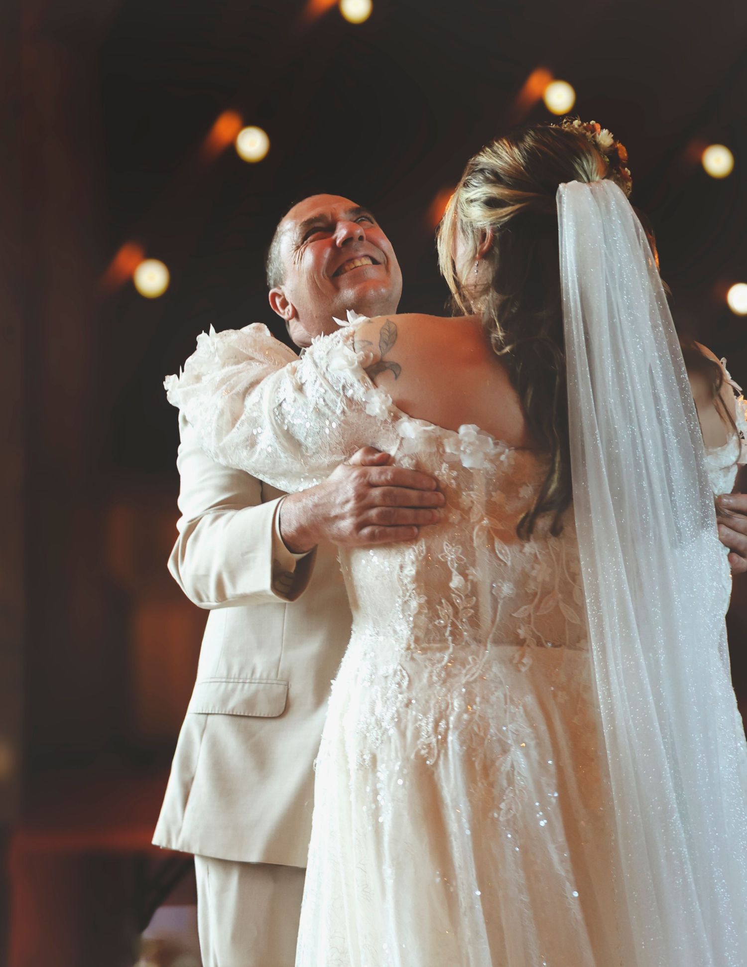 Father and bride sharing emotional hug during wedding dance, bride wearing lace gown and veil.