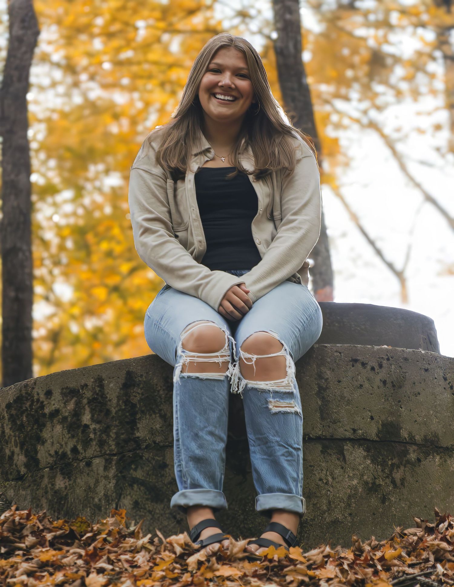 Smiling young woman in ripped jeans sitting on stone wall surrounded by autumn foliage.