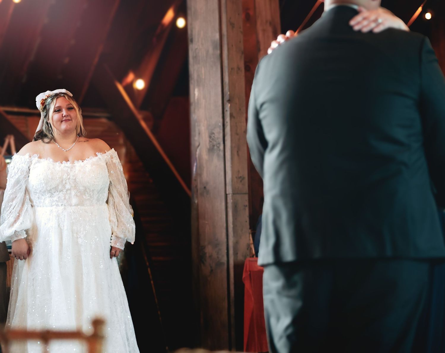 Bride in off-shoulder lace wedding gown shares first dance with groom in dark suit inside rustic barn venue.