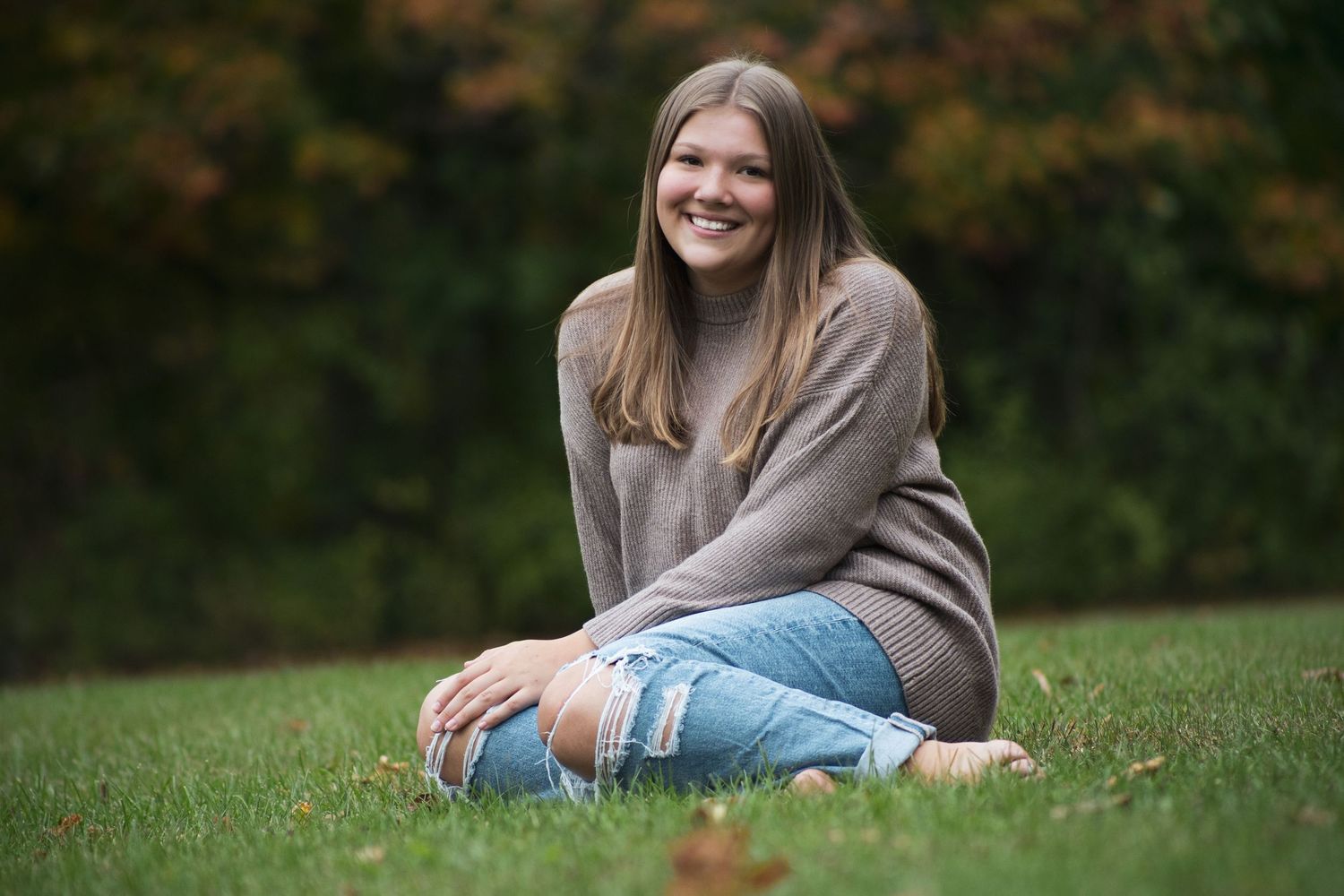 Young woman smiling, sitting cross-legged on grass in ripped jeans and brown sweater, with autumn trees behind her.