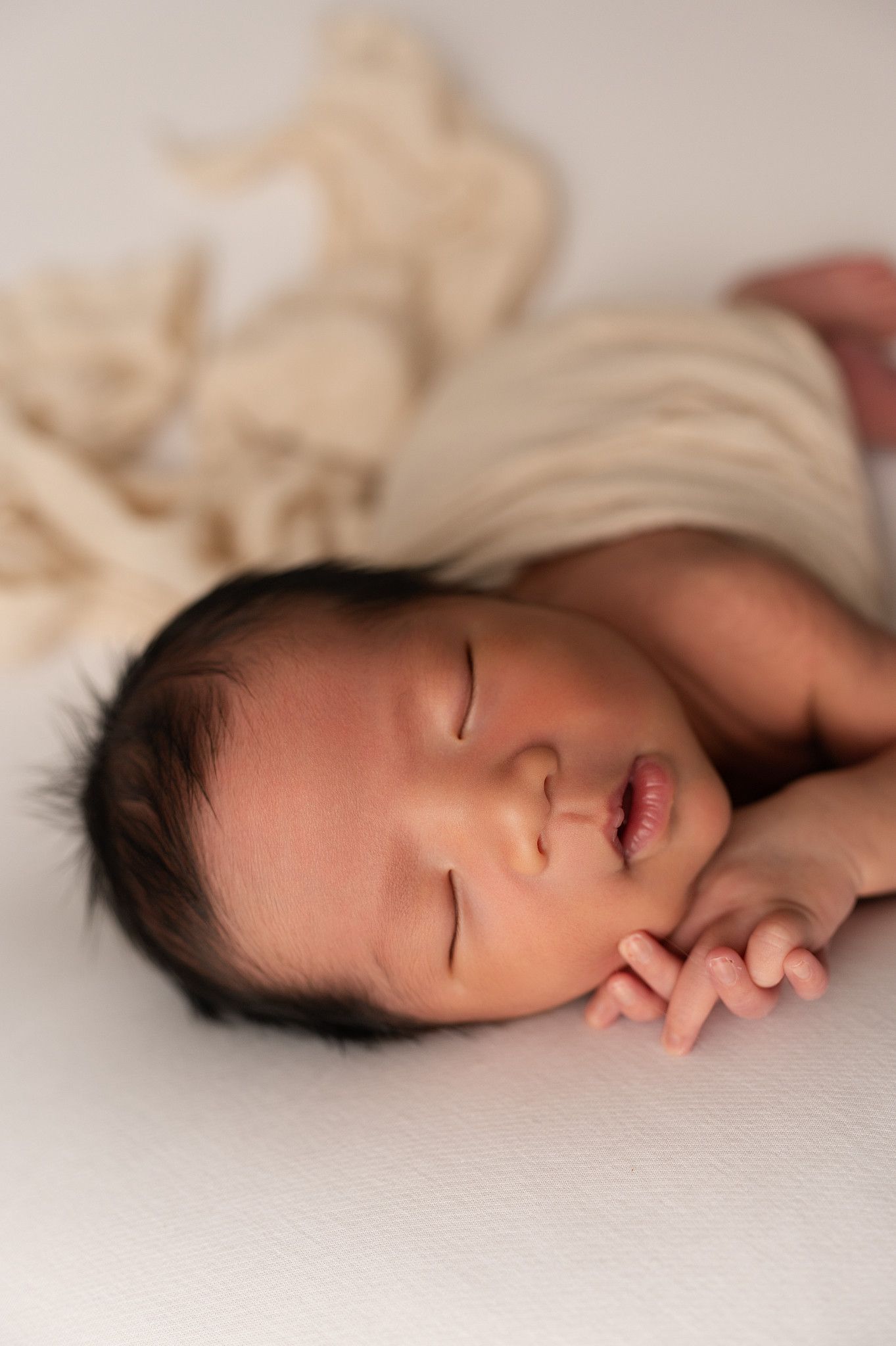 Close-up portrait of sleeping newborn baby with dark hair resting on hands with cream wrap behind.