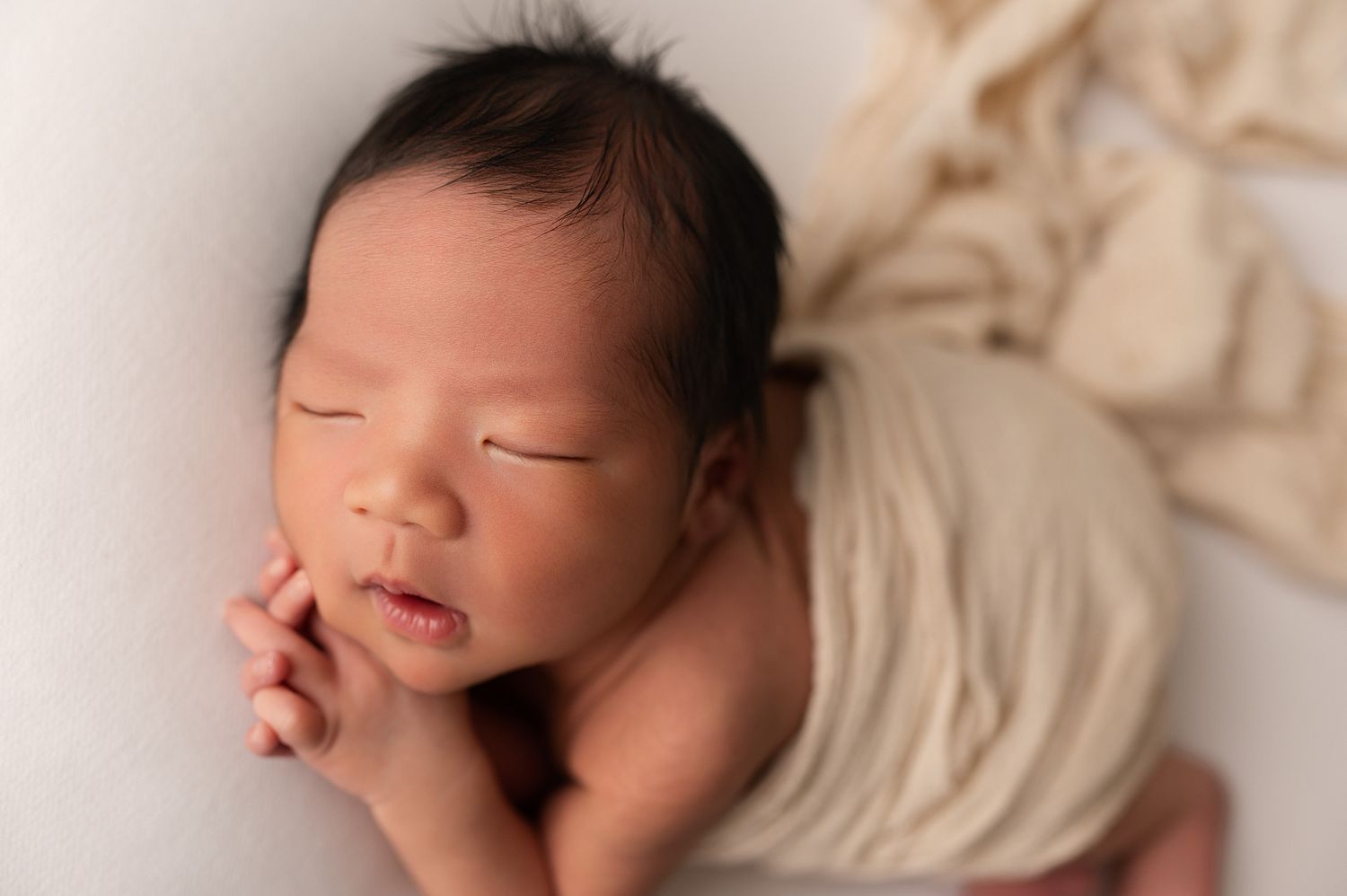 Sleeping newborn baby with dark hair posed on white surface wrapped in soft cream swaddle cloth.