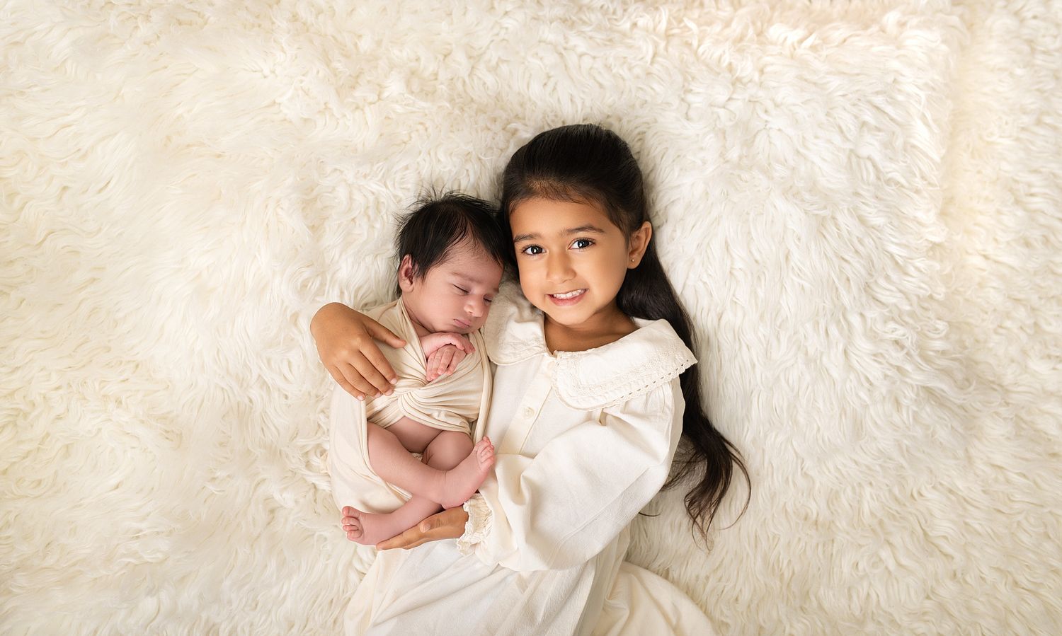 Older sister in white dress smiling while holding newborn baby on fluffy cream fur rug in professional photo.