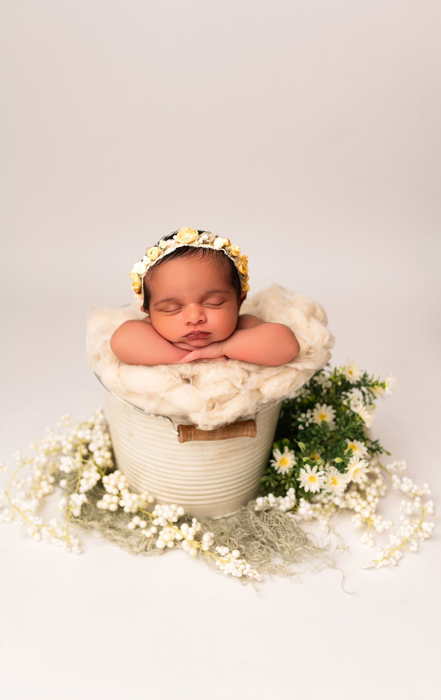 Sleeping newborn baby in a rustic bucket with soft fur, wearing a daisy headband, surrounded by white flowers.