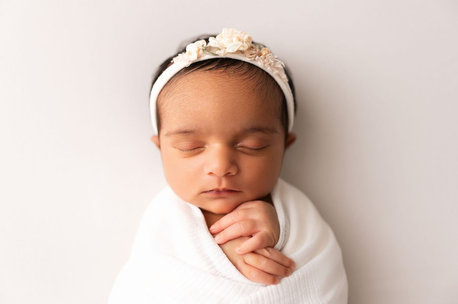 Close-up of a sleeping newborn in a white swaddle wearing a delicate floral headband, hands tucked under chin.