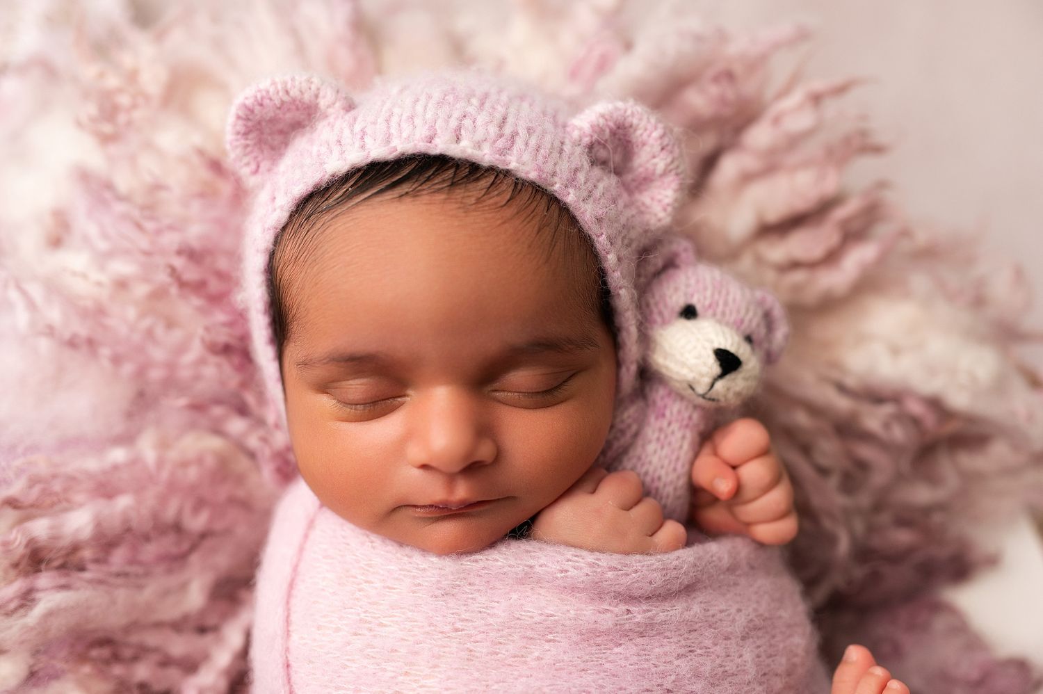Newborn baby sleeping in pink bear bonnet holding a tiny knit teddy bear on a fluffy pink backdrop.