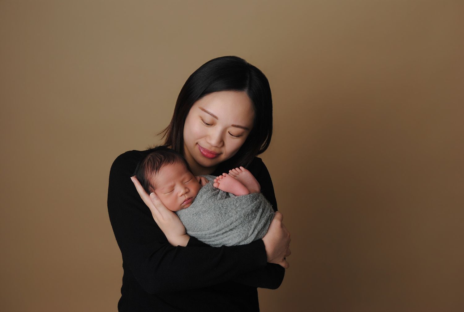 Asian mother in black outfit tenderly holding sleeping swaddled newborn baby on tan background.