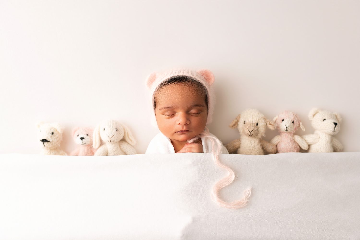 Newborn baby sleeping in white wrap wearing pink bear bonnet surrounded by small stuffed animal toys.