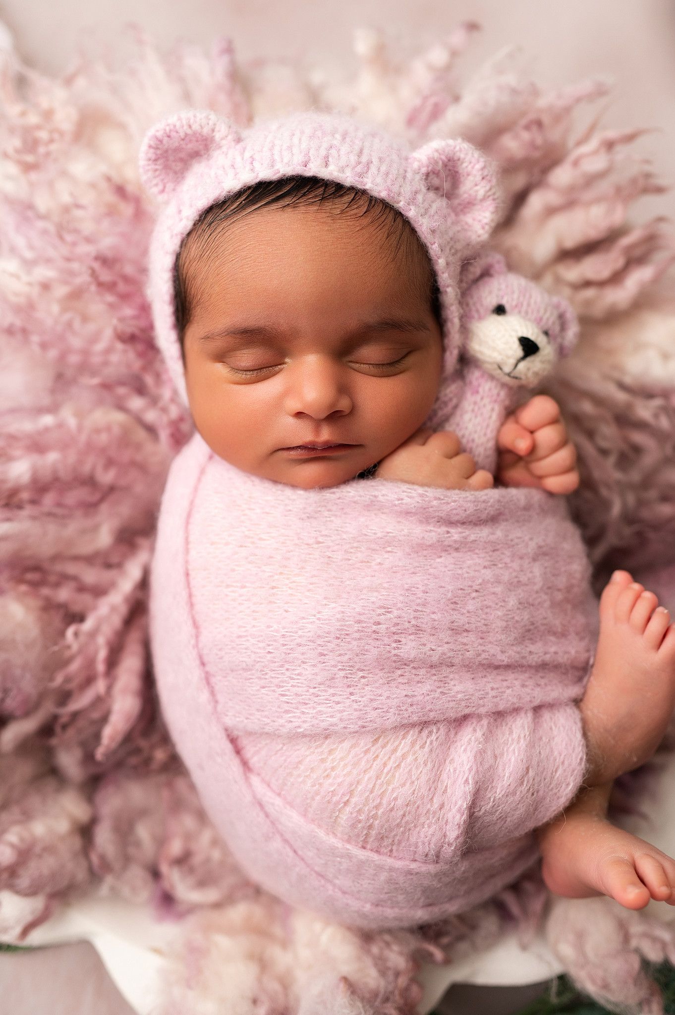 Pink-swaddled newborn wearing a bear ear bonnet, holding a small knit teddy bear on a floral pink blanket.