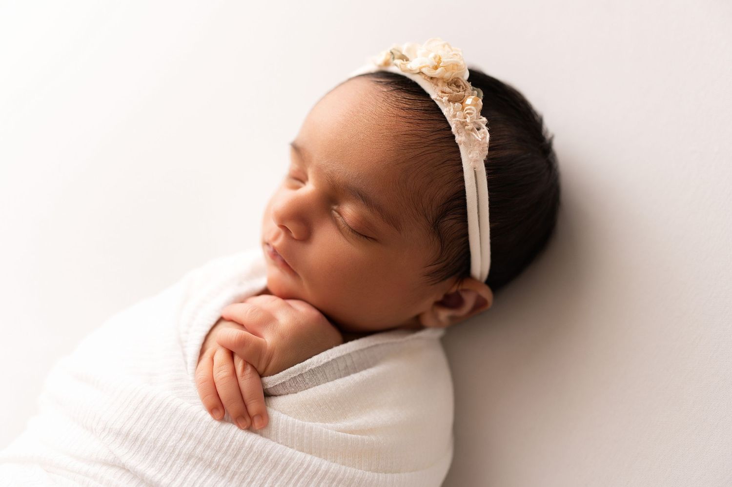 Peaceful sleeping newborn swaddled in white wrap wearing a delicate floral headband on a white background.