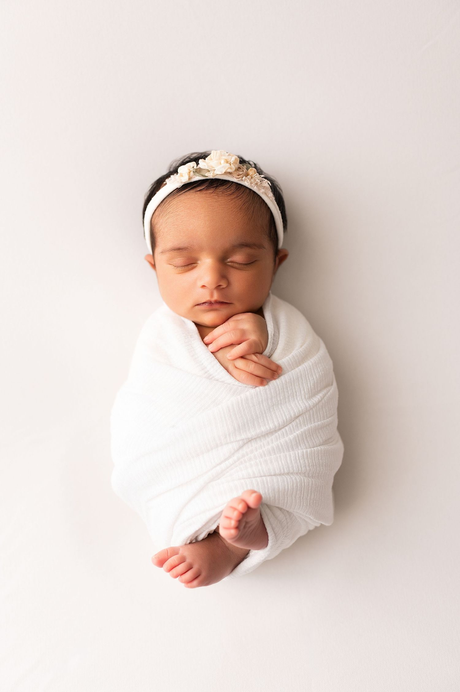 Sleeping newborn wrapped in white swaddle with a floral headband, tiny foot peeking out on white backdrop.