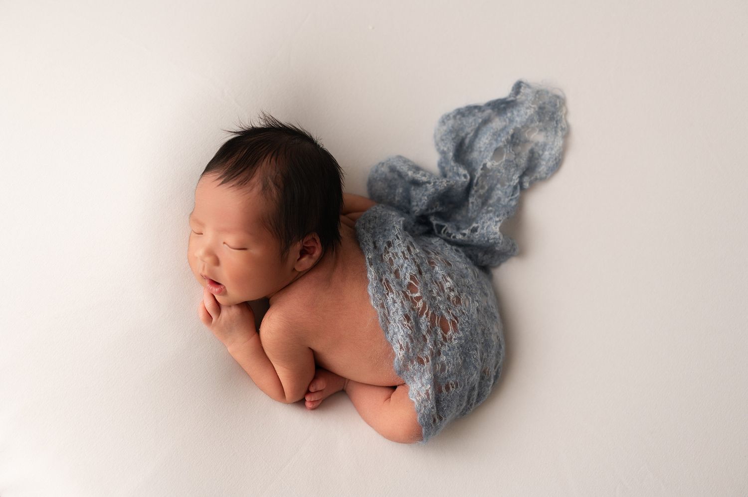 Sleeping newborn baby posed on white surface draped in flowing blue lace wrap during professional photo session.