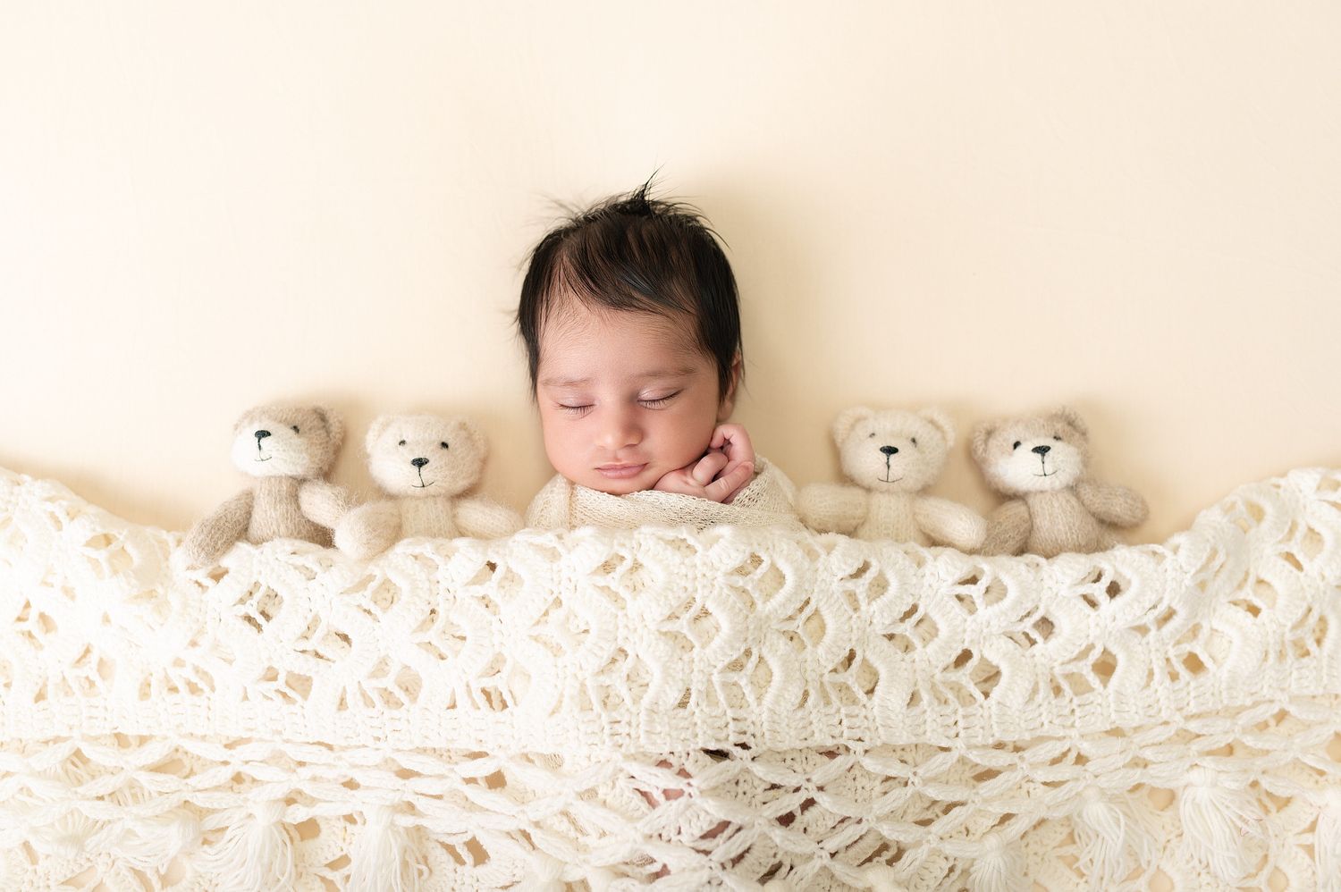 Sleeping newborn baby tucked under a cream knit blanket surrounded by four small teddy bears on a beige background.