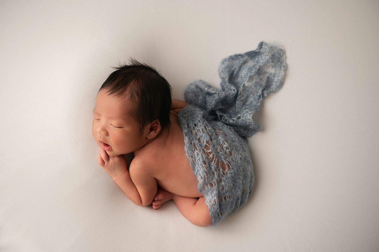 Sleeping newborn baby posed with blue lace wrap trailing elegantly on soft white background.