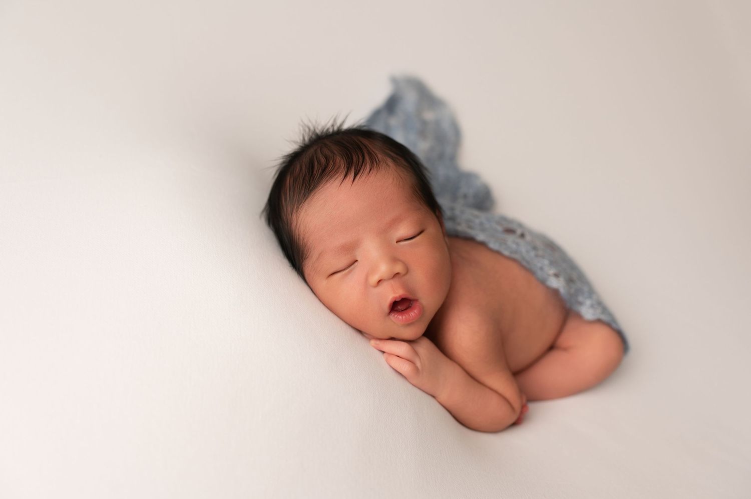 Peaceful sleeping newborn baby wrapped in blue cloth lying on white background with mouth slightly open.