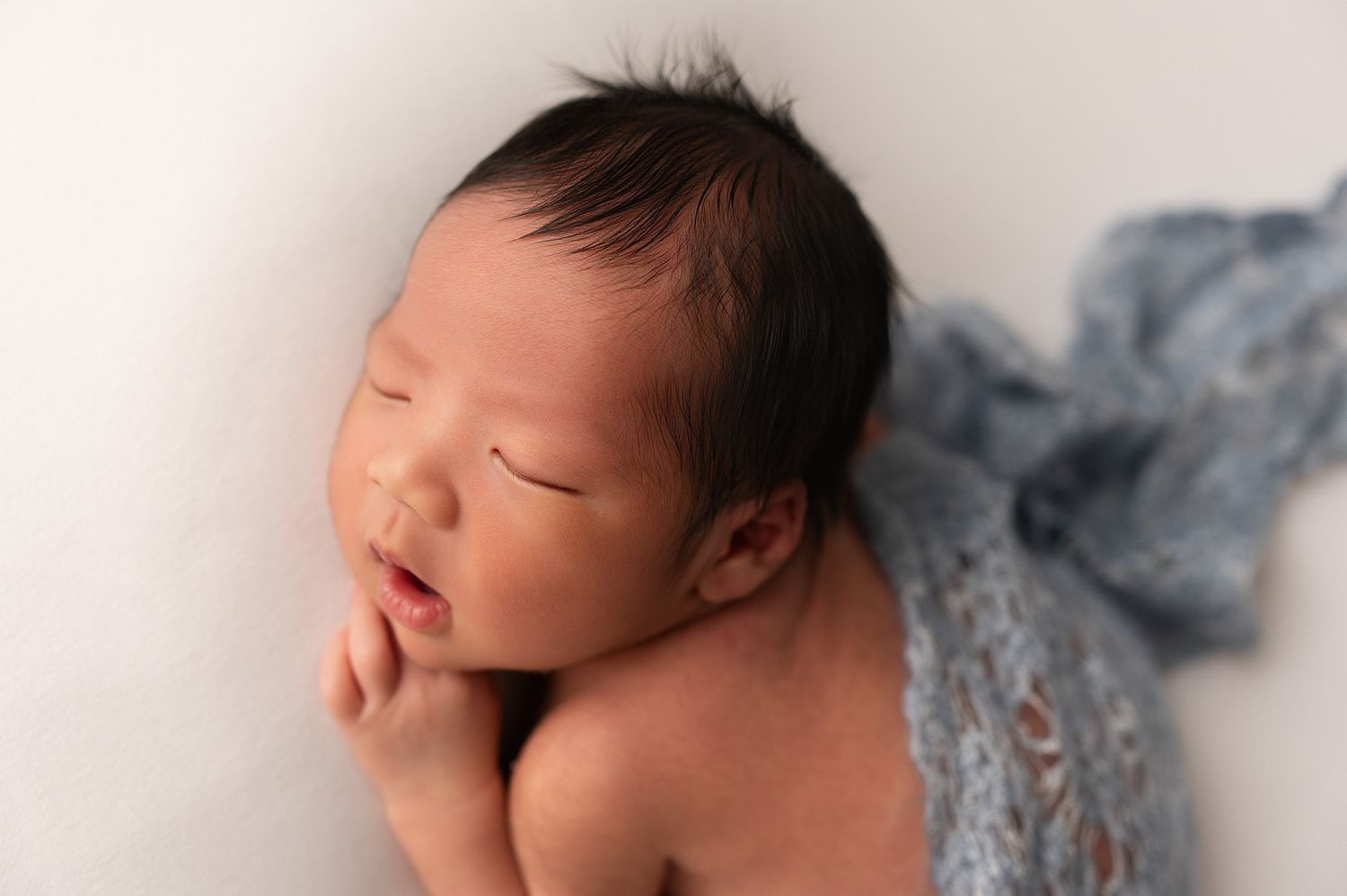 Close-up of sleeping newborn baby with dark hair, resting chin on hands with blue wrap on white background.