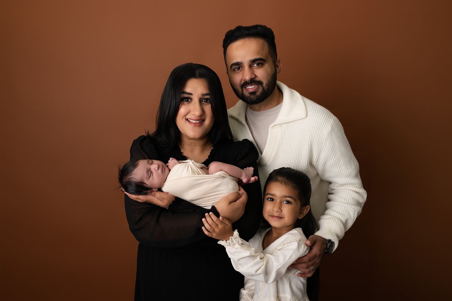 Family portrait with parents and two children, newborn baby, and young girl, posing against a warm brown background.