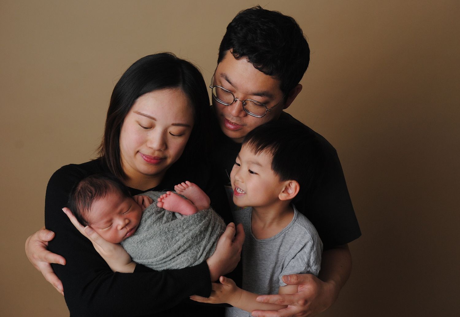 Happy family of four with newborn baby and young son posing on tan background.