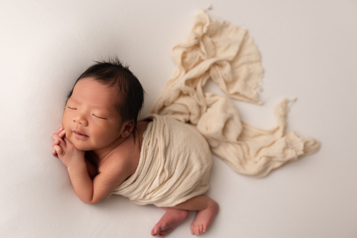 Newborn baby sleeping peacefully wrapped in cream cloth with fabric trailing on white background.