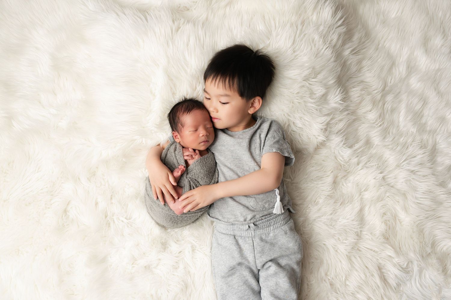 Young boy lying on white fur rug gently holding sleeping newborn baby sibling in soft gray wrap.