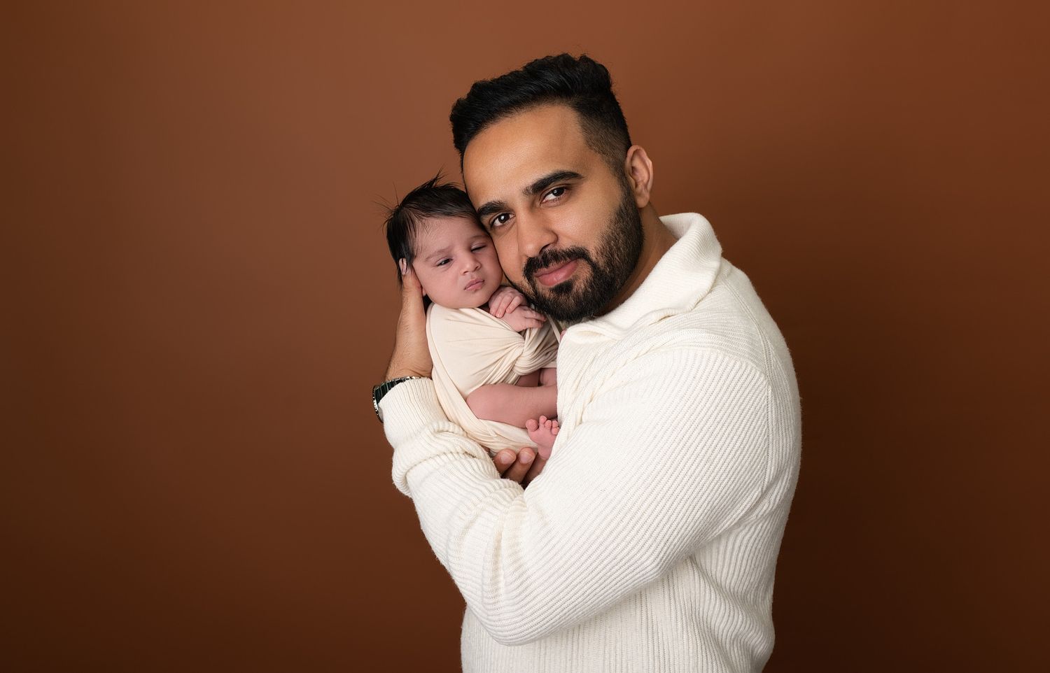 A man in a white sweater tenderly holds a sleeping newborn baby against a warm brown studio background.