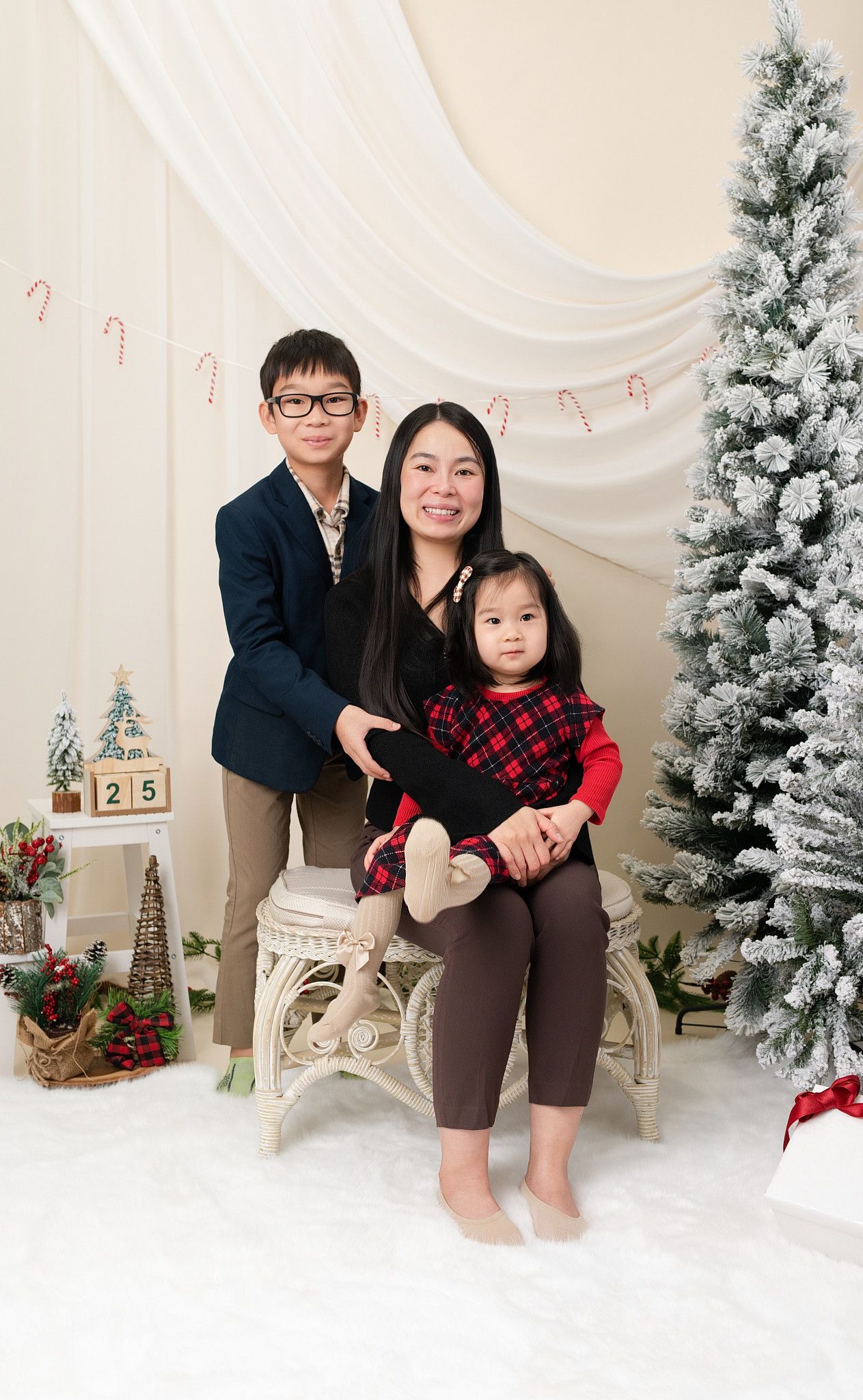 A smiling woman sits with a boy in glasses and a young girl in a plaid dress in a Christmas studio with snowy trees and festive decor.