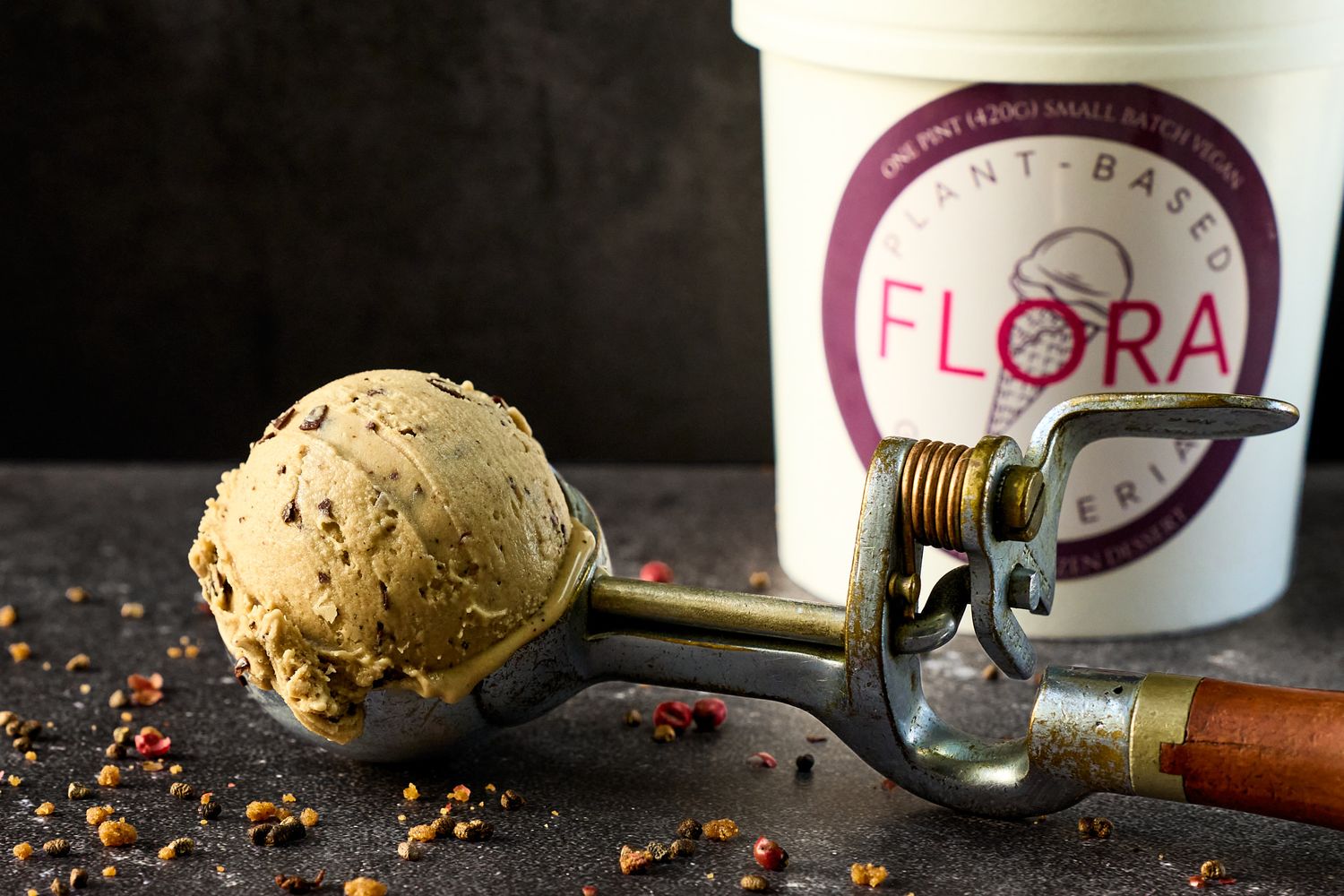 Plant-based Flora ice cream scoop on a metal scooper beside the container on dark surface.
