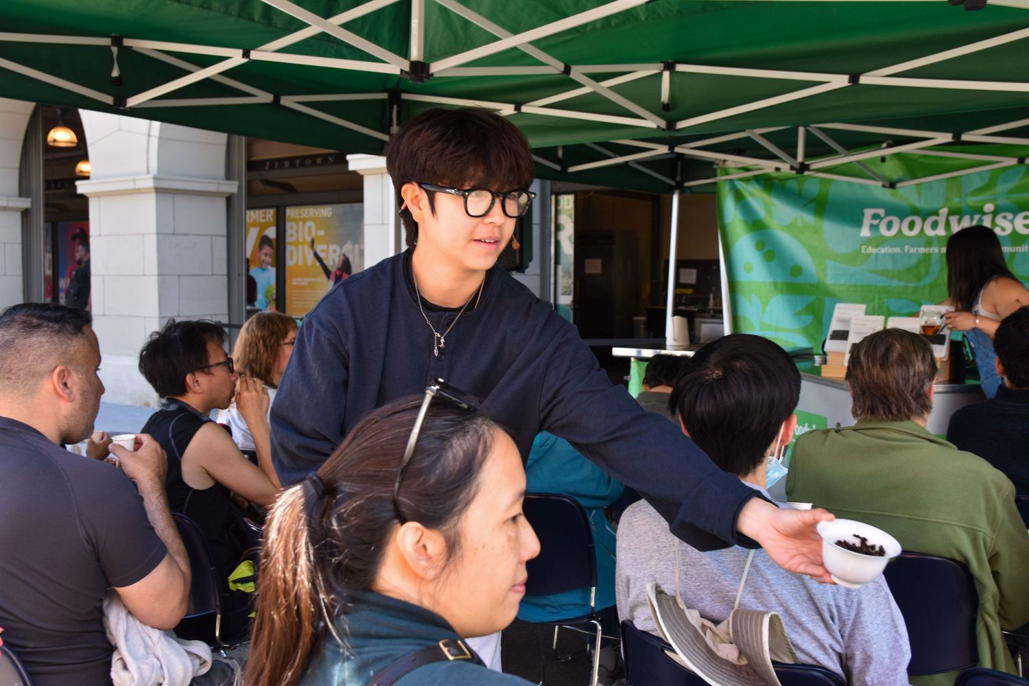 A young man in glasses serves food samples to attendees seated at an outdoor Foodwise market event.
