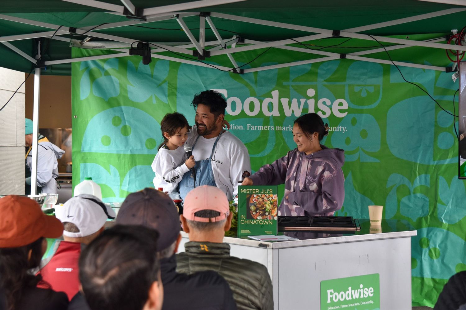 Two people present a cooking demonstration at a Foodwise farmers market education booth outdoors.