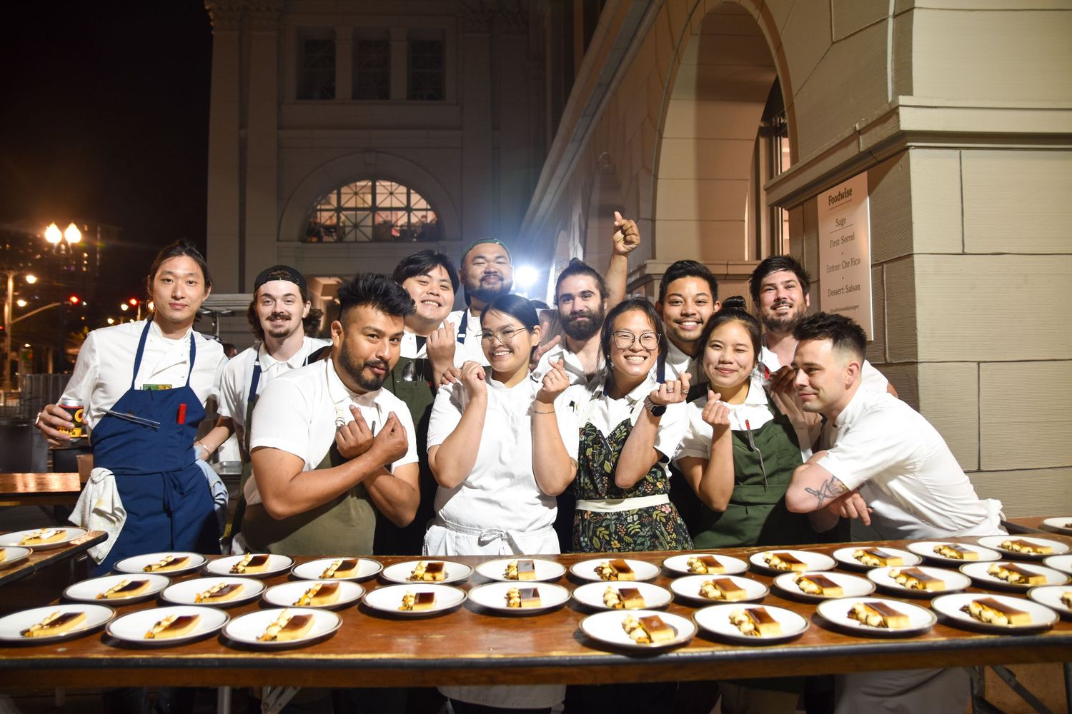 A large group of chefs and staff pose together smiling behind a table of plated dishes at night.