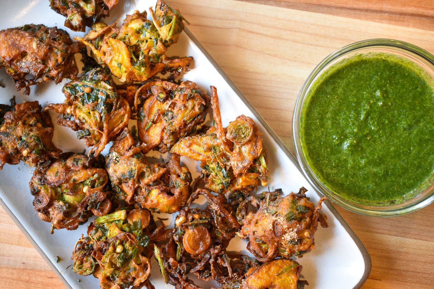 Crispy spiced vegetable fritters on a white platter served with a vibrant green herb dipping sauce.