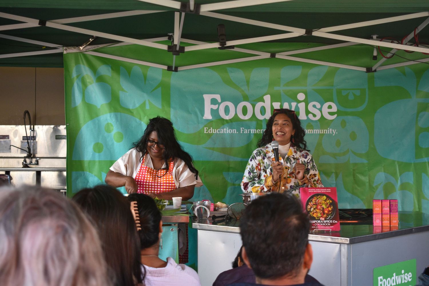 Two women present a live cooking demonstration under a Foodwise tent at an outdoor farmers market.