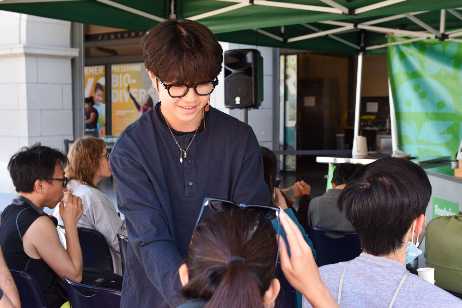 A young man with glasses smiles while handing out samples to seated guests at an outdoor food event.