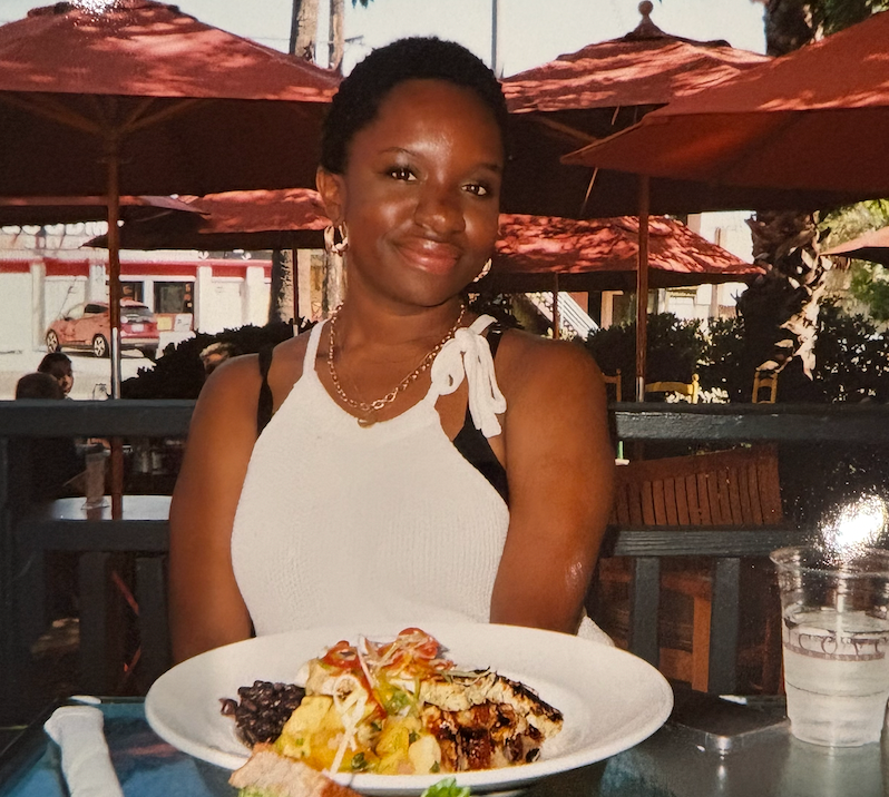 A smiling woman in a white dress poses with a colorful plate of food at an outdoor restaurant patio.