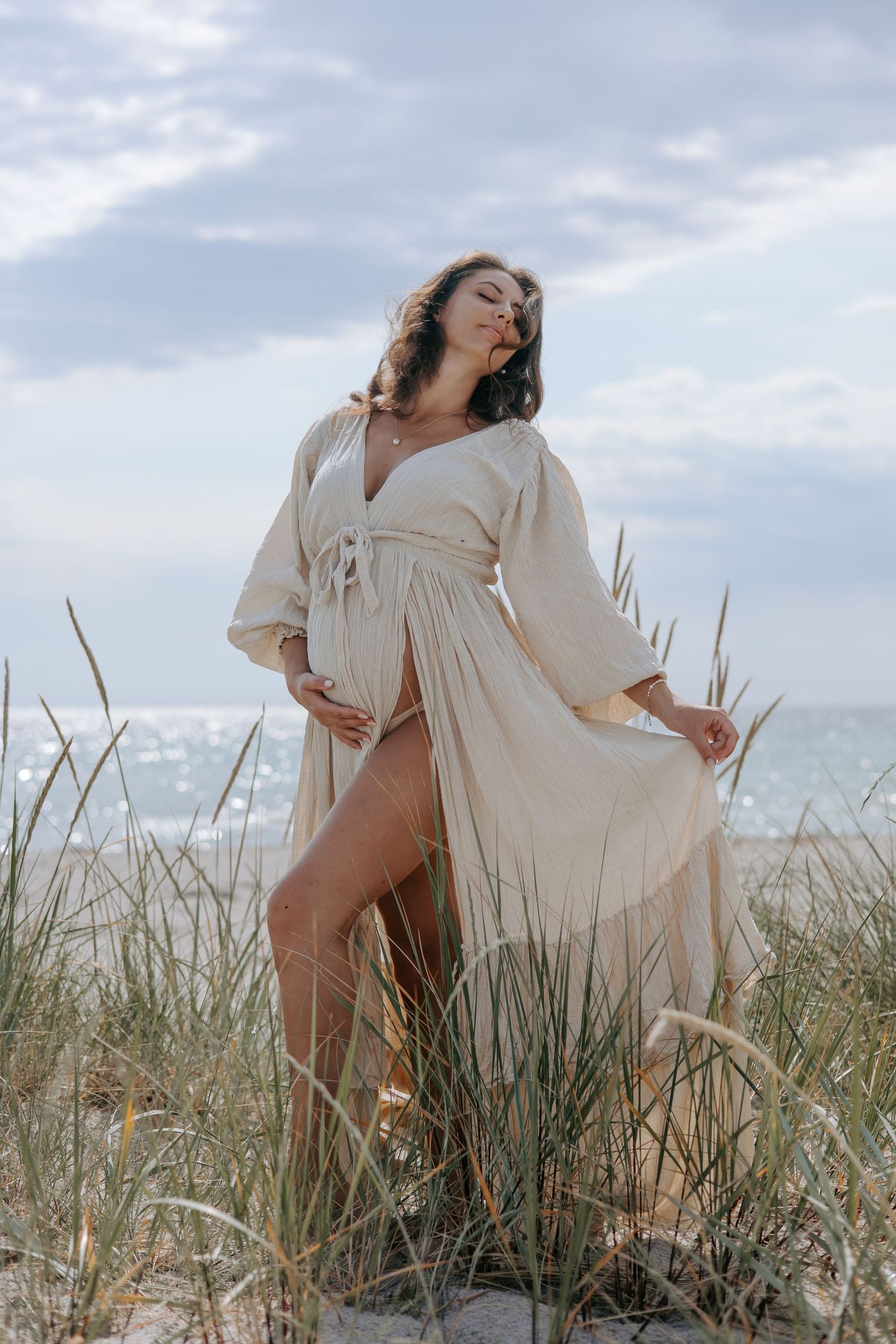 Pregnant woman in flowing beige wrap dress standing among beach grass with ocean backdrop.