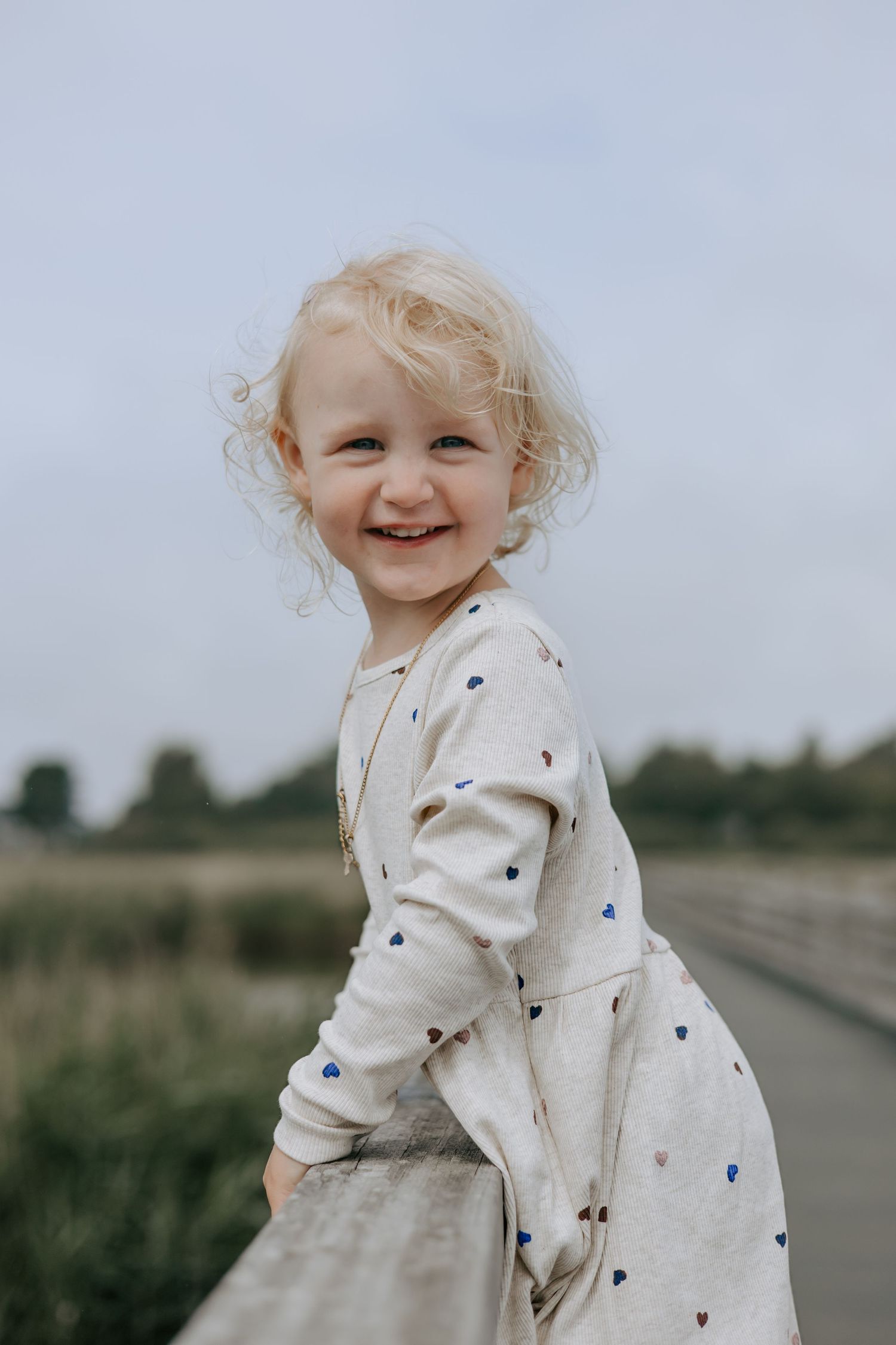Happy blonde toddler girl smiling in a heart-print dress outdoors.