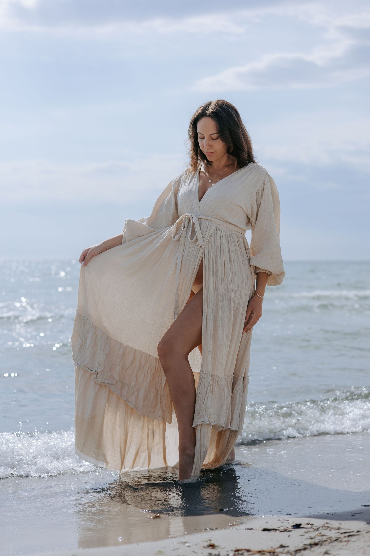 Pregnant woman in flowing cream dress standing in ocean waves on sandy beach.