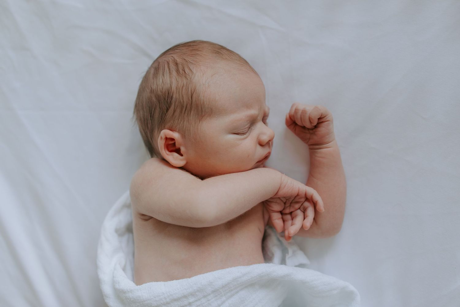 Peaceful newborn baby sleeping wrapped in white swaddle blanket on white surface.