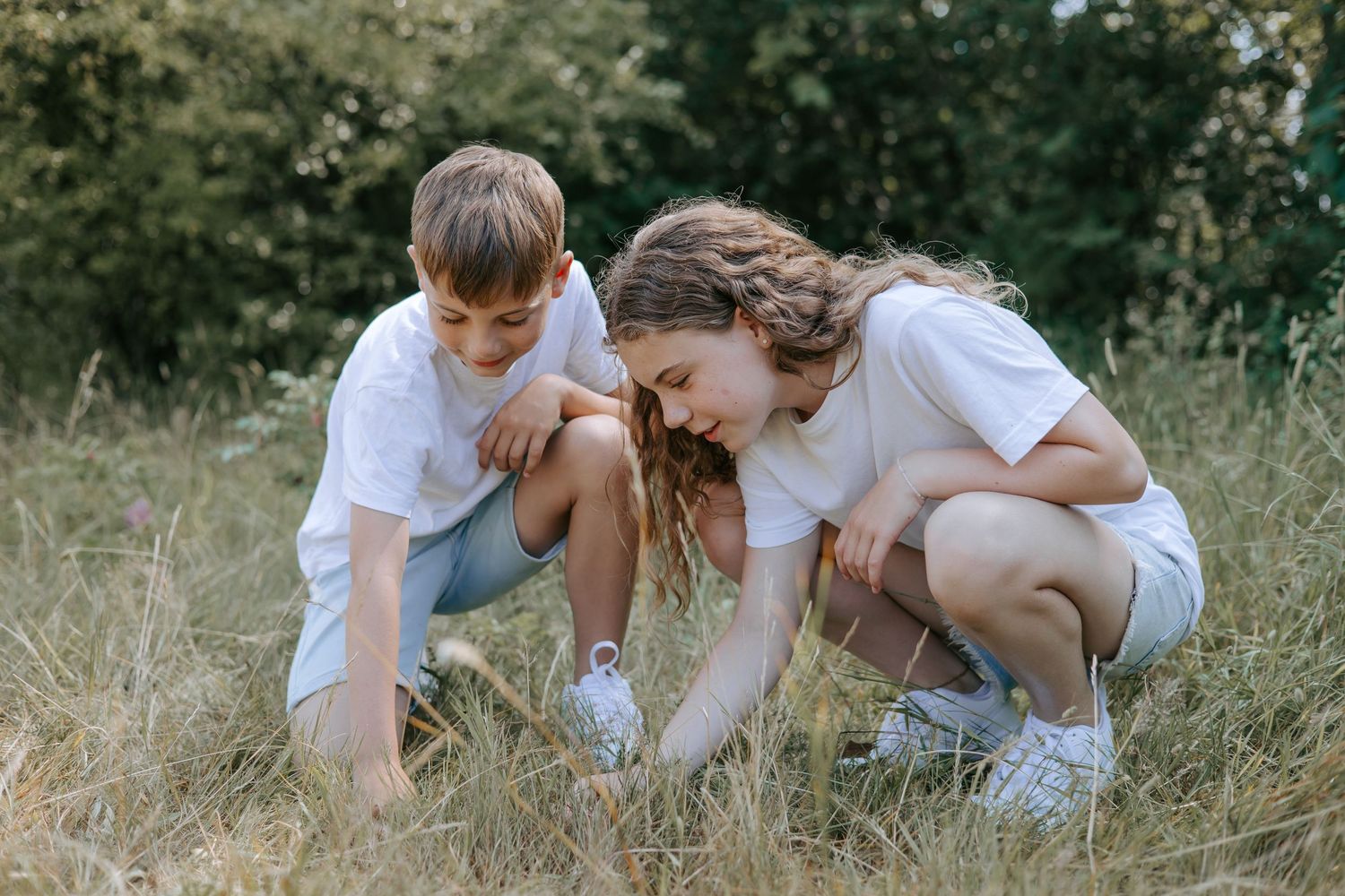 Brother and sister crouching together in a grassy field outdoors.