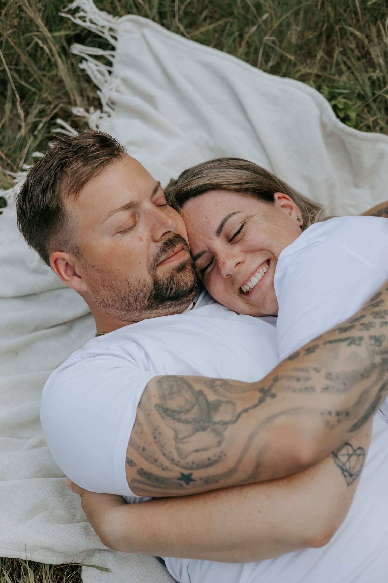 Couple lying on a blanket in a field, embracing and smiling, man with tattoos in white t-shirts.