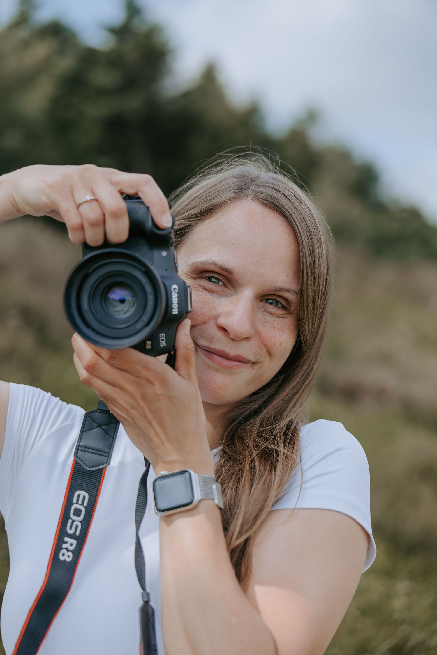 A smiling woman photographer holding a Canon EOS R8 camera outdoors in nature.