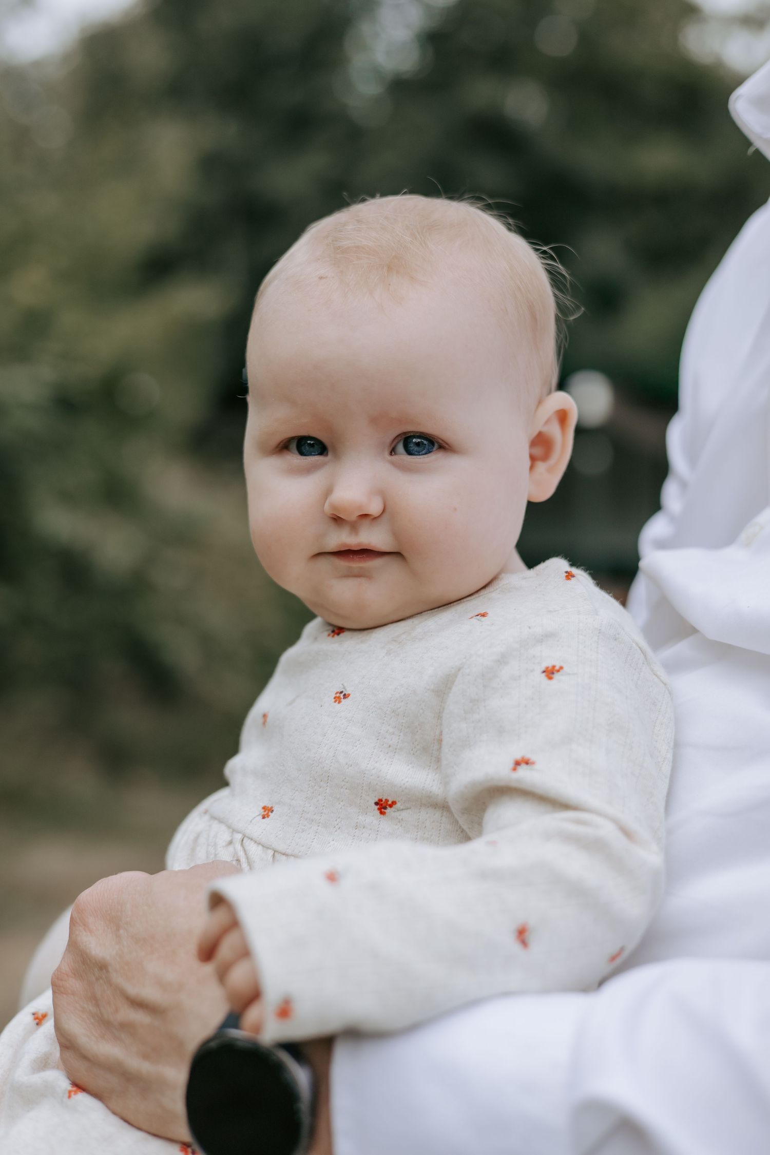 Adorable blue-eyed baby in white floral outfit being held outdoors with green trees behind.